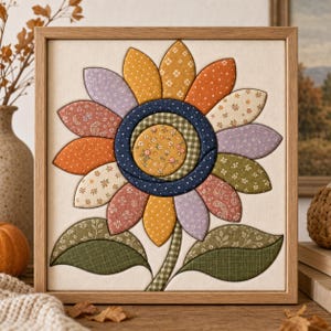 May include: A square framed fabric wall art piece featuring a central sunflower motif. The flower has a yellow patterned center, a dark blue ring, and multi-colored fabric petals in shades of orange, yellow, lavender, and cream. Two green fabric leaves are at the base.