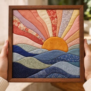 Puede incluir: Una pieza de arte mural de tela cuadrada en un marco de madera oscura con un diseño de rayos de sol acolchado. La escena muestra un sol amarillo dorado elevándose sobre paneles de tela ondulados en tonos azules, verdes y crema con diversos patrones florales.