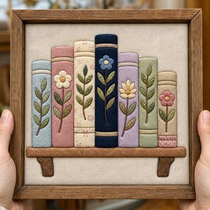 May include: A square wooden frame containing a fabric wall hanging of seven books on a shelf. The books are in shades of blue, pink, cream, navy, lavender, and green, each featuring embroidered floral stems and leaves on their spines.