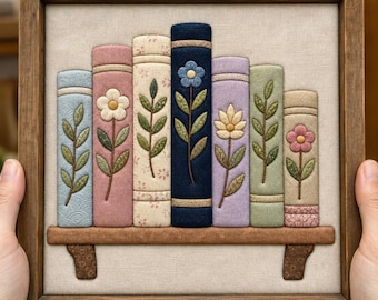 May include: A square wooden frame containing a fabric wall hanging of seven books on a shelf. The books are in shades of blue, pink, cream, navy, lavender, and green, each featuring embroidered floral stems and leaves on their spines.