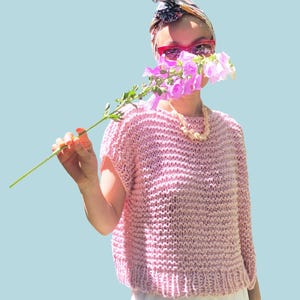 May include: A light pink, short-sleeved knit top with a textured pattern. The word "PATTERN" is in a white rectangle at the top. The top is paired with white pants. A person is holding a flower in front of their face.