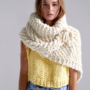 May include: A woman wearing a white knitted cowl neck scarf over a yellow knitted sleeveless top and blue jeans.