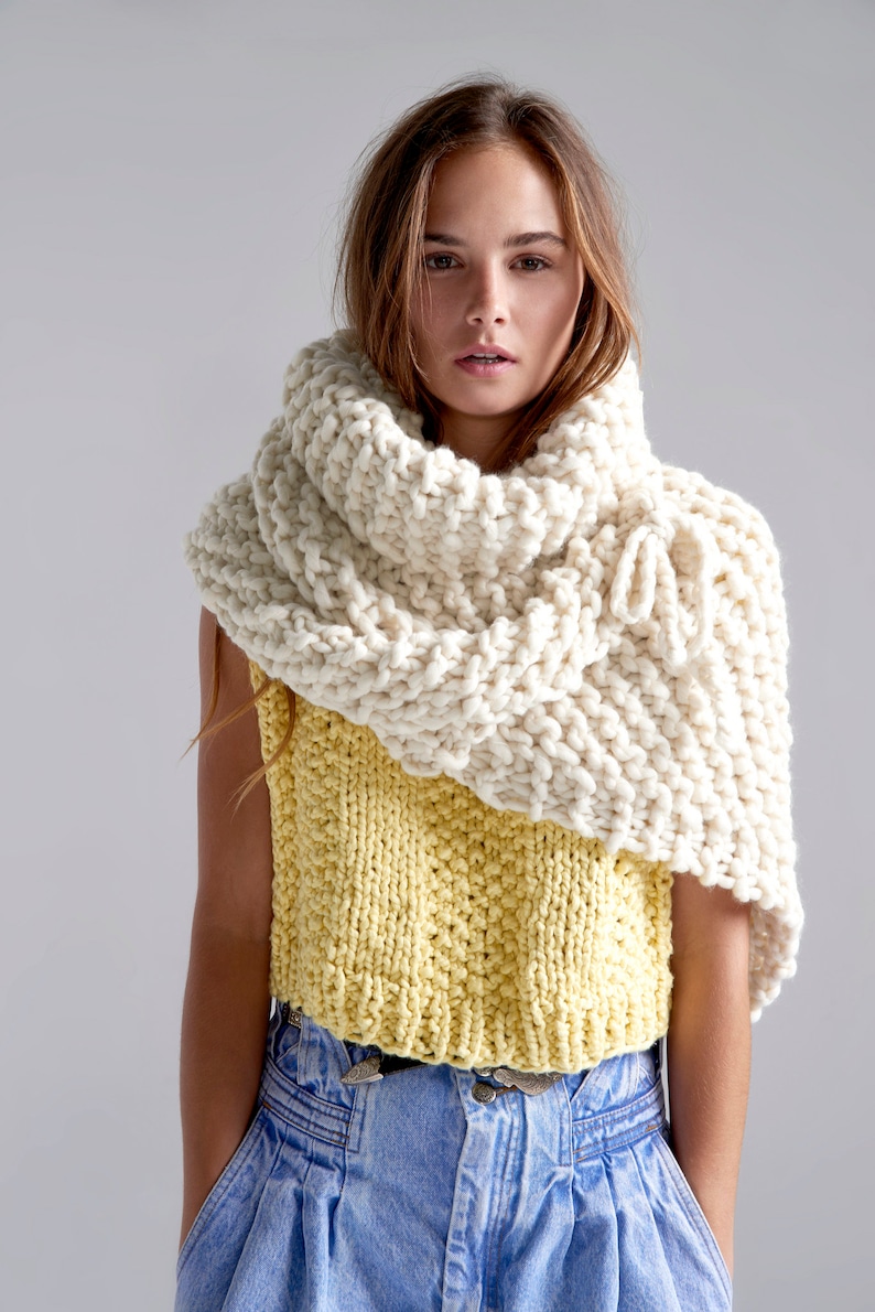 May include: A woman wearing a white knitted cowl neck scarf over a yellow knitted sleeveless top and blue jeans.