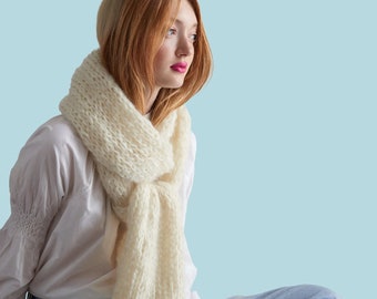 May include: A woman wearing a white shirt and blue jeans with a white knitted scarf around her neck. The scarf is loosely draped around her neck and shoulders. The woman is looking off to the side and has red hair.