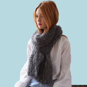 May include: A woman wearing a white shirt and blue jeans is wearing a gray knitted scarf. The scarf is thick and fluffy, and it is draped around her neck. The word "PATTERN" is written in white letters on a light blue background.