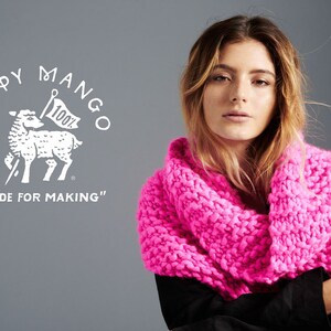May include: A woman wearing a bright pink chunky knit scarf. The scarf is made of thick yarn and has a loose, open weave. The woman is wearing a black shirt and has long brown hair. The background is a gray wall. The image also includes a white logo with a sheep and the text "LOOPY MANGO" and "MADE FOR MAKING".