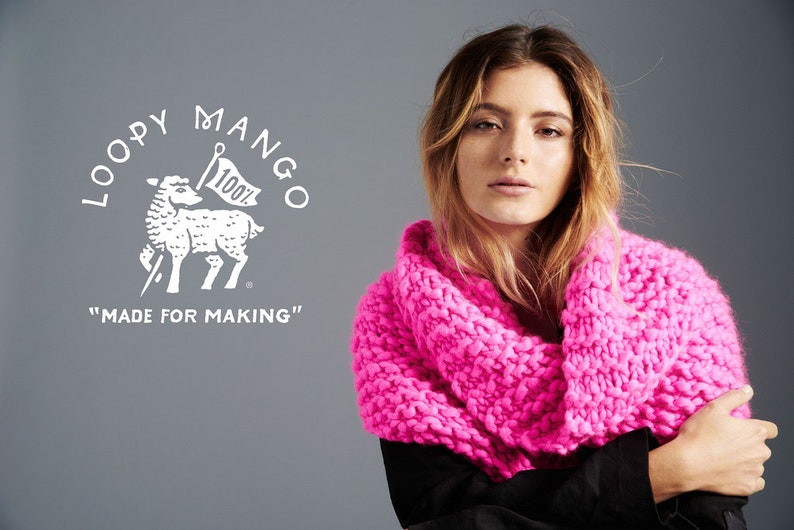 May include: A woman wearing a bright pink chunky knit scarf. The scarf is made of thick yarn and has a loose, open weave. The woman is wearing a black shirt and has long brown hair. The background is a gray wall. The image also includes a white logo with a sheep and the text "LOOPY MANGO" and "MADE FOR MAKING".