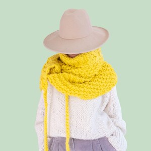 May include: A DIY KIT label in pink and white text above a beige felt hat, a chunky knit bright yellow scarf with long dangling ends, a white cable knit sweater, and light purple corduroy trousers. The items are layered to form an outfit.