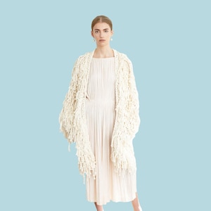 May include: A woman in a cream dress and a fringed, off-white shawl. The word "PATTERN" is in a white rectangle. She wears blue shoes with gold details, against a light blue backdrop. The outfit is elegant.