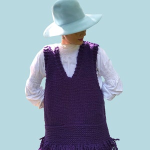 May include: A purple knitted vest with a fringe hem. The vest has a V-neck and is worn over a white shirt. The person is wearing a light blue hat.