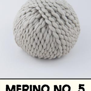 May include: A round ball of light gray, thick, twisted yarn. Below the yarn ball, a rectangular label with a cream-colored background features the black text MERINO NO. 5 in a bold, sans-serif font. The yarn has a chunky, braided texture.