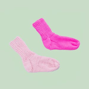 May include: A DIY KIT featuring two hand-knitted socks, one in bright neon pink and one in soft pastel pink. Both socks have a ribbed cuff and a standard heel construction. The text DIY KIT appears at the top and INTERMEDIATE at the bottom.