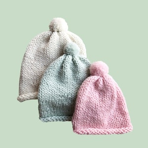 May include: A DIY KIT for an ABSOLUTE BEGINNER featuring three hand-knitted beanies with pom-poms. The hats are arranged in a cluster, colored in cream, light sage green, and soft pastel pink, each featuring a thick, chunky knit texture.