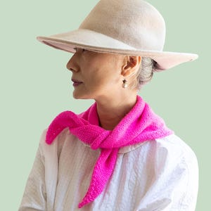 May include: A DIY KIT for a bright hot pink knitted neckerchief, featuring a textured stitch pattern. The kit includes materials to create this accessory, which is shown tied around the neck, paired with a beige felt hat and a white shirt.