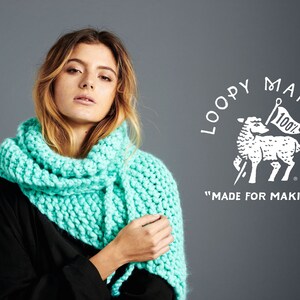 May include: A woman wearing a teal blue knitted scarf. The scarf is made with a chunky yarn and has a textured stitch. The background is a gray wall with a white logo that says "Loopy Mango" and "Made for Making".