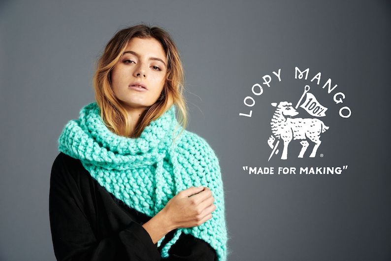 May include: A woman wearing a teal blue knitted scarf. The scarf is made with a chunky yarn and has a textured stitch. The background is a gray wall with a white logo that says "Loopy Mango" and "Made for Making".