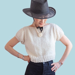 May include: A woman wearing a black hat and a cream-colored knitted top with short sleeves. The word "PATTERN" is in white text at the top. She is also wearing dark pants and jewelry.