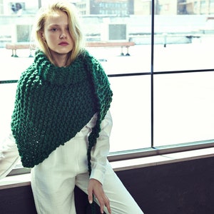 May include: A woman wearing a white jumpsuit with a large, chunky green knitted scarf. The scarf is draped around her neck and shoulders, and the ends are hanging down her back. She is standing in front of a window with a view of a city skyline.