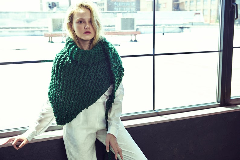 May include: A woman wearing a white jumpsuit with a large, chunky green knitted scarf. The scarf is draped around her neck and shoulders, and the ends are hanging down her back. She is standing in front of a window with a view of a city skyline.