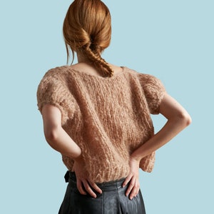 Puede incluir: Una mujer lleva un suéter de punto marrón claro de corte holgado y mangas cortas. Lleva pantalones de cuero negro y tiene el pelo recogido en una trenza.