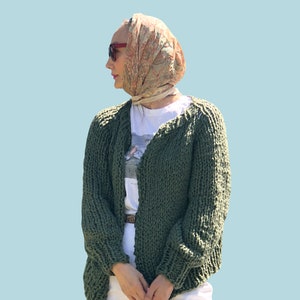 May include: A green knitted cardigan with long sleeves and a v-neck. The cardigan is open and the person wearing it is holding a pink bag.
