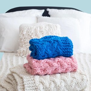 Op de afbeelding: Een stapel van drie dikke gebreide dekens in blauw, roze en crème. De dekens zijn netjes opgevouwen en op een bed met witte kussens gelegd. Het woord "PATTERN" staat in een wit kader bovenaan.