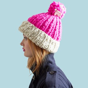 Op de afbeelding: Een roze en witte gebreide beanie met een pompon bovenop. De beanie wordt gedragen door een persoon met lang rood haar die een donkerblauwe jas draagt.