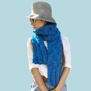 Op de afbeelding: Een vrouw draagt een grijze hoed, zonnebril, een wit shirt en een dikke blauwe gebreide sjaal. Het woord "PATTERN" staat in een witte rechthoek bovenaan de afbeelding. De sjaal heeft een levendige blauwe kleur.