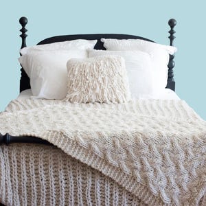 Op de afbeelding: Een bed met een zwart frame, wit beddengoed en een crèmekleurige gebreide deken. Een decoratief kussen met franjes ligt op het bed. Het woord "PATTERN" staat in een witte rechthoek boven het bed.