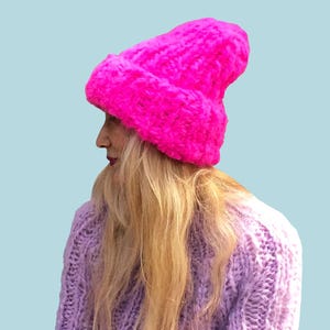 Op de afbeelding: Een felroze gebreide muts met een getextureerde uitstraling wordt gedragen door een persoon met lang blond haar. De persoon draagt ook een lichtpaarse gebreide trui. Het woord "PATTERN" staat in een witte rechthoek bovenaan de afbeelding.