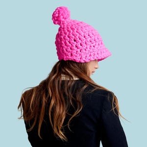 Op de afbeelding: Een felroze, dik gebreide muts met een afgeronde kroon en een kleine pompon. De muts wordt gedragen door een persoon met lang bruin haar, tegen een lichtblauwe achtergrond. Het woord "PATTERN" wordt in witte tekst bovenaan weergegeven.