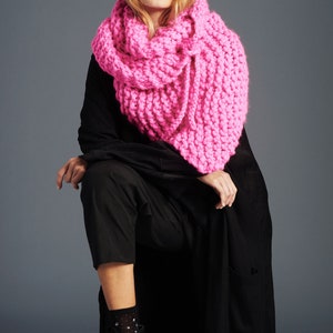 May include: A woman wearing a black long-sleeved dress with a bright pink chunky knit scarf. She is sitting on a wooden box and wearing black ankle boots with white patterned socks.