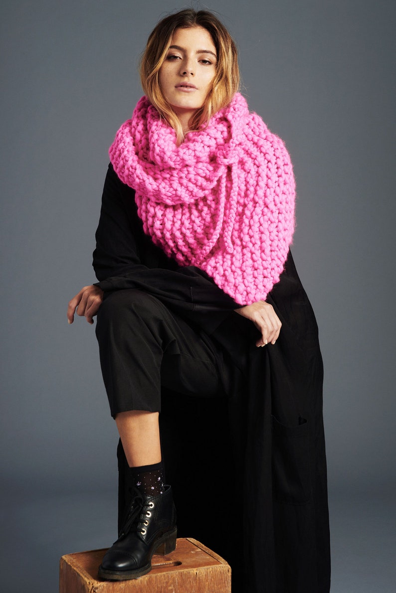 May include: A woman wearing a black long-sleeved dress with a bright pink chunky knit scarf. She is sitting on a wooden box and wearing black ankle boots with white patterned socks.