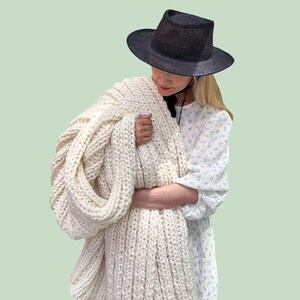 May include: A DIY KIT for a chunky knit blanket featuring thick, cream-colored yarn in a ribbed pattern. The kit is labeled BEGINNER and includes the materials to create this large, textured throw blanket with a soft, heavy appearance.