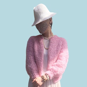 May include: A pink knitted cardigan with a white hat and a floral patterned face mask. The cardigan has a loose fit and long sleeves.