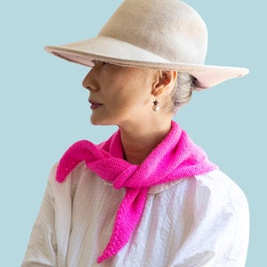 Op de afbeelding: Een beige vilten hoed en een fel neonroze gebreide driehoekige sjaal die om de nek is geknoopt. De afbeelding bevat de tekst PATTERN bovenaan en BEGINNER onderaan, wat duidt op een breiproject voor mensen aan het begin van hun niveau.