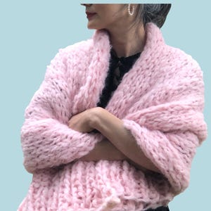 May include: A light pink, chunky knit shawl. The shawl is wrapped around the shoulders and arms. The word "PATTERN" is in white block letters at the top of the image. The shawl is made of a thick, soft yarn and has a textured appearance.