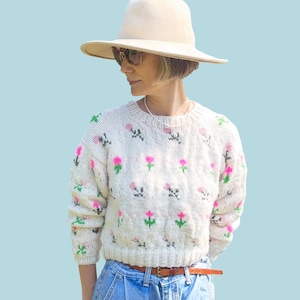 May include: A cream-colored sweater with pink and green floral embroidery. The sweater is paired with light blue jeans and a brown belt. A wide-brimmed hat and sunglasses are worn. The word "PATTERN" is in a white rectangle at the top.