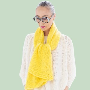 May include: A bright yellow chunky knit scarf worn over a white open-knit sweater. The image features the text DIY KIT at the top and BEGINNER at the bottom. The scarf has a thick, textured yarn construction with a simple loop knot style.