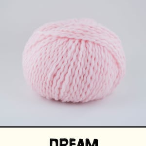 May include: A round ball of light pink yarn with a thick, twisted texture. Below the yarn, a horizontal rectangular banner contains the word DREAM in bold, black, sans-serif capital letters against a pale cream-colored background.