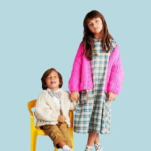 Op de afbeelding: Twee kinderen in stijlvolle outfits staan tegen een lichtblauwe achtergrond. Eén kind draagt een crèmekleurig vest, een bruine broek en een vlinderdas. Het andere kind draagt een geruite jurk met een felroze trui. Het woord "PATTERN" staat bovenaan.