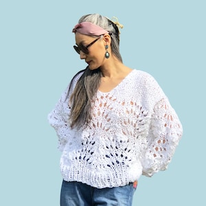 Könnte beinhalten: Ein weißer Pullover mit offenem Gewebe und V-Ausschnitt. Der Pullover hat ein Blumenmuster und lange Ärmel. Das Wort "PATTERN" befindet sich in einem weißen Rechteck oben im Bild. Die Person trägt eine blaue Jeans.