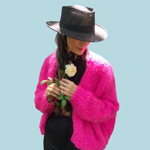 May include: A woman wearing a black hat and a bright pink fuzzy cardigan holds a single white rose.