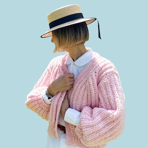 May include: A light pink, chunky knit cardigan with a V-neck and long sleeves. The cardigan is worn over a white collared shirt and white pants. A straw boater hat with a black band and ribbon is worn. The word "PATTERN" is in white text at the top.