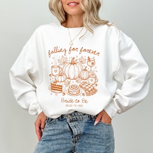 May include: A white crewneck sweatshirt featuring a brown graphic print of pumpkins, autumn leaves, a slice of cake, a cinnamon roll, a plaid blanket, and mugs. The text reads "falling for forever", "Bride to Be", and "DALLAS • TX • 2026".