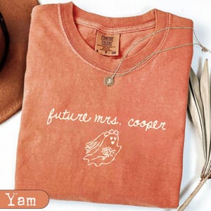May include: A terracotta-colored short-sleeve t-shirt with a crew neckline and a Comfort Colors brand label. The shirt features white text that reads future mrs. cooper above a line drawing of a ghost wearing a veil and holding a bouquet.