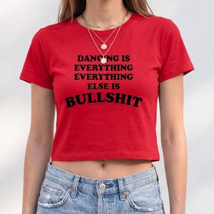 May include: A bright red short-sleeved crop top featuring the black printed text DANCING IS EVERYTHING EVERYTHING EVERYTHING ELSE IS BULLSHIT centered on the chest. The garment is paired with light blue denim jeans and a layered gold necklace.