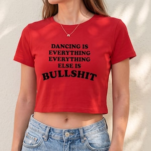 Könnte beinhalten: Ein leuchtend rotes, kurzärmeliges bauchfreies Oberteil mit dem schwarzen Aufdruck DANCING IS EVERYTHING EVERYTHING EVERYTHING ELSE IS BULLSHIT mittig auf der Brust. Das Kleidungsstück hat einen Rundhalsausschnitt und wird mit einer hellblauen Jeans kombiniert.