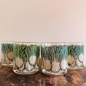 May include: A set of four clear glass tumblers featuring a repeating graphic print of white onions with green tops. Each glass has a cylindrical shape with a smooth rim and a flat base, showcasing a detailed botanical illustration in black ink.