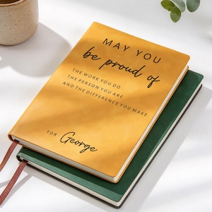 Może przedstawiać: Musztardowy notatnik leżący na ciemnozielonym notatniku. Żółta okładka zawiera czarny tekst: MAY YOU be proud of THE WORK YOU DO THE PERSON YOU ARE AND THE DIFFERENCE YOU MAKE FOR George. Dwie cienkie wstążki pełniące funkcję zakładek wystają z boku.